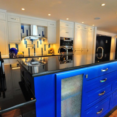 A white tall wall of custom cabinets and pantries is a great backdrop to seventeen-feet of electric blue, gloss island storage. The island with custom designed cookbook and drawer storage is capped with black gloss granite. Channel lighting installed into the granite washes the island and chrome hardware with light. 